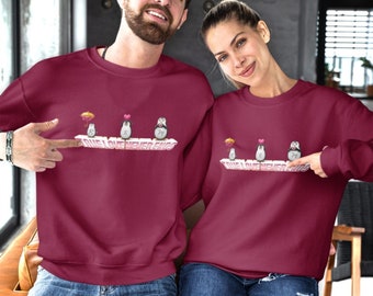 May include: Two people wearing maroon sweatshirts with a graphic of penguins holding umbrellas and hearts with the text "True Love Never Ends".
