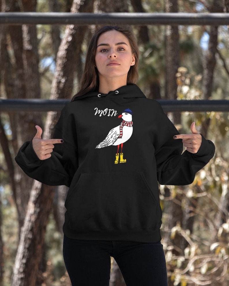May include: Black hooded sweatshirt with a white seagull wearing a blue and red striped scarf and a blue hat. The seagull is wearing yellow rain boots. The text "Moin" is printed on the sweatshirt.