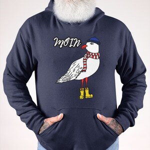 May include: A navy blue hoodie with a white seagull wearing a blue hat, red and white scarf, and yellow rain boots. The word "Moin" is printed in white above the seagull.