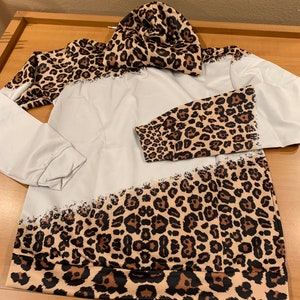 May include: A white hoodie with a leopard print design on the sleeves and bottom hem. The hoodie has a hood and a drawstring.