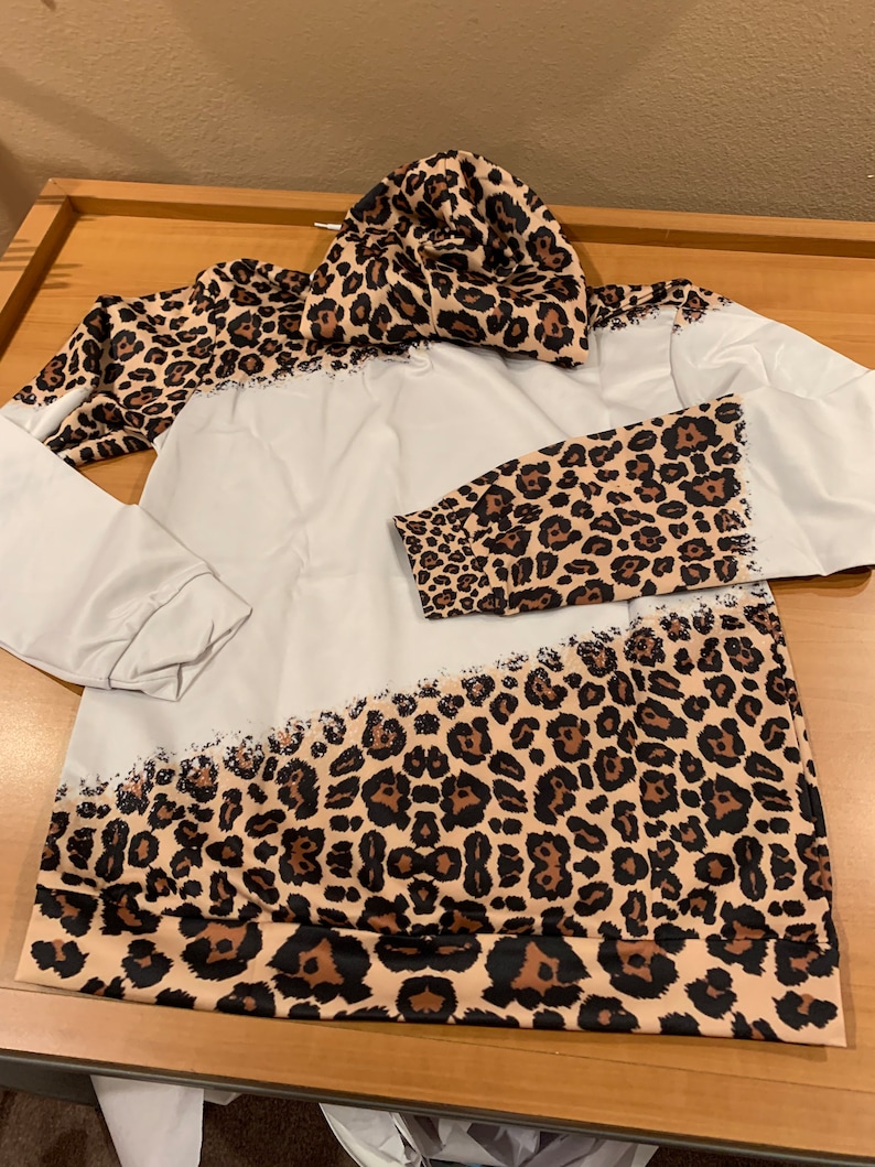 May include: A white hoodie with a leopard print design on the sleeves and bottom hem. The hoodie has a hood and a drawstring.
