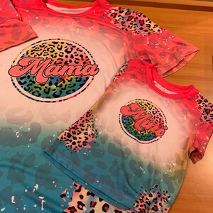 May include: Two t-shirts with a tie-dye effect and a leopard print design. The larger shirt has the word "Mama" in a rainbow circle. The smaller shirt has the word "Mini" in a rainbow circle.