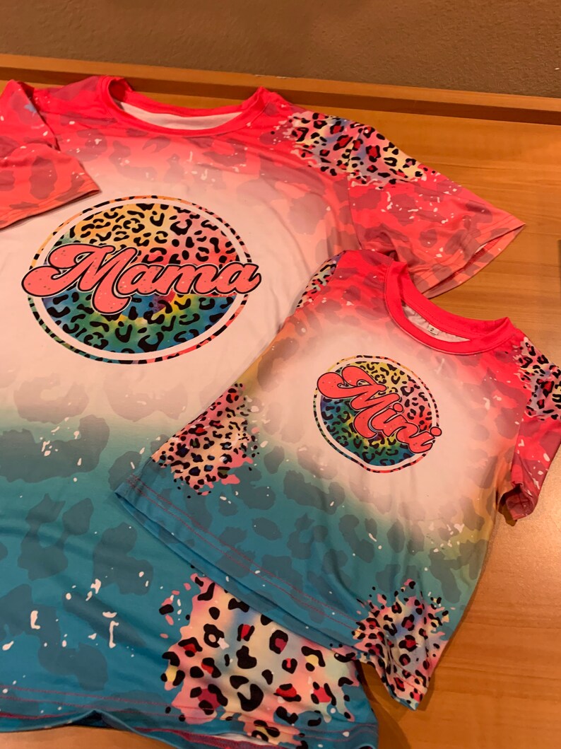 May include: Two t-shirts with a tie-dye effect and a leopard print design. The larger shirt has the word "Mama" in a rainbow circle. The smaller shirt has the word "Mini" in a rainbow circle.
