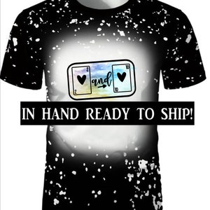 May include: Black t-shirt with a white splatter paint design. The shirt features a graphic of two playing cards with hearts, the numbers 2 and 10, and the word "and" with an arrow pointing to the right.