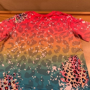 May include: A colorful t-shirt with a rainbow ombre design and leopard print. The shirt has a relaxed fit and short sleeves.