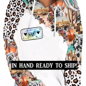 May include: White hoodie with a colorful print of a cow, cactus, and a cowboy hat on the sleeves. The front of the hoodie has a graphic of two playing cards with hearts and the word "and" in between. The text "IN HAND READY TO SHIP!" is printed in black at the bottom of the hoodie.