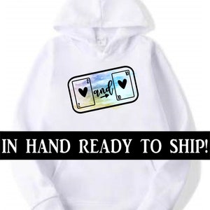 May include: White hoodie with a graphic of two playing cards, a 2 of hearts and a 10 of hearts, with an arrow pointing from the 2 to the 10 and the word "and" written above the arrow. The graphic is on a watercolor background.