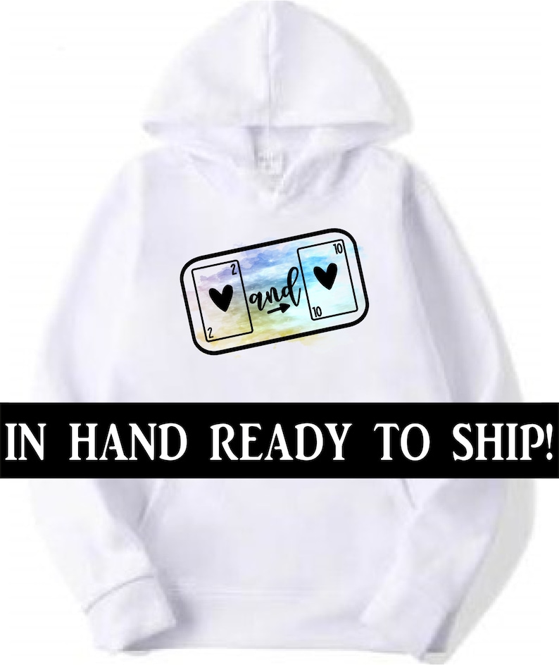 May include: White hoodie with a graphic of two playing cards, a 2 of hearts and a 10 of hearts, with an arrow pointing from the 2 to the 10 and the word "and" written above the arrow. The graphic is on a watercolor background.