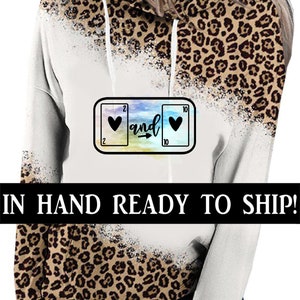 May include: White and brown leopard print hoodie with a colorful graphic of two playing cards with hearts and the word "and" in between. The text "IN HAND READY TO SHIP!" is at the bottom of the image.