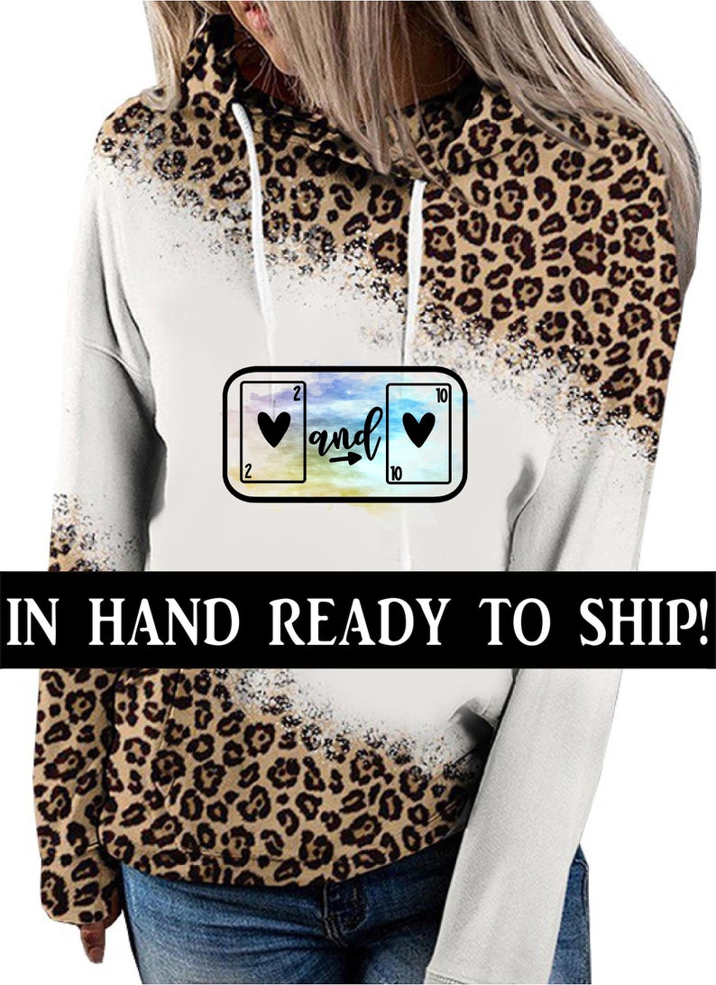 May include: White and brown leopard print hoodie with a colorful graphic of two playing cards with hearts and the word "and" in between. The text "IN HAND READY TO SHIP!" is at the bottom of the image.