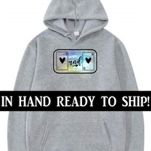 May include: A light gray hoodie with a front pocket and a hood. The design on the front features two playing cards with hearts and the word "and" in a script font. The text "IN HAND READY TO SHIP!" is across the bottom.