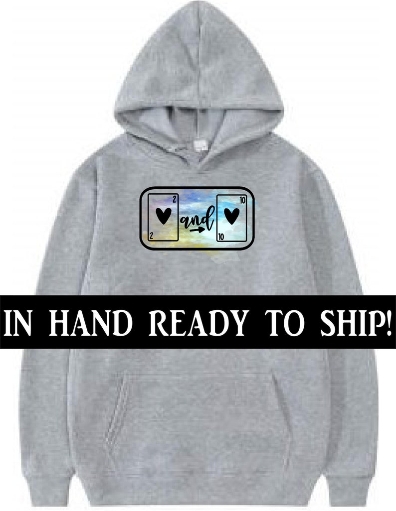 May include: A light gray hoodie with a front pocket and a hood. The design on the front features two playing cards with hearts and the word "and" in a script font. The text "IN HAND READY TO SHIP!" is across the bottom.