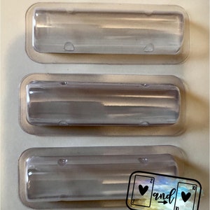 May include: Three clear, rectangular plastic containers with rounded edges are stacked on a light beige surface. Each container has a slightly curved shape and small, raised details. A small sticker with the word "and" and playing card symbols is in the bottom right corner.