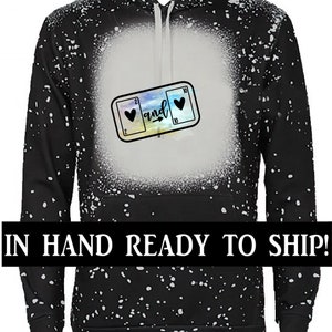 K&ouml;nnte beinhalten: Schwarzer Hoodie mit einem wei&szlig;en Spritzmuster. Der Hoodie hat eine Grafik mit zwei Spielkarten mit Herzen, den Zahlen 2 und 10 und dem Wort "and" dazwischen. Der Text "IN HAND READY TO SHIP!" befindet sich am unteren Rand des Bildes.