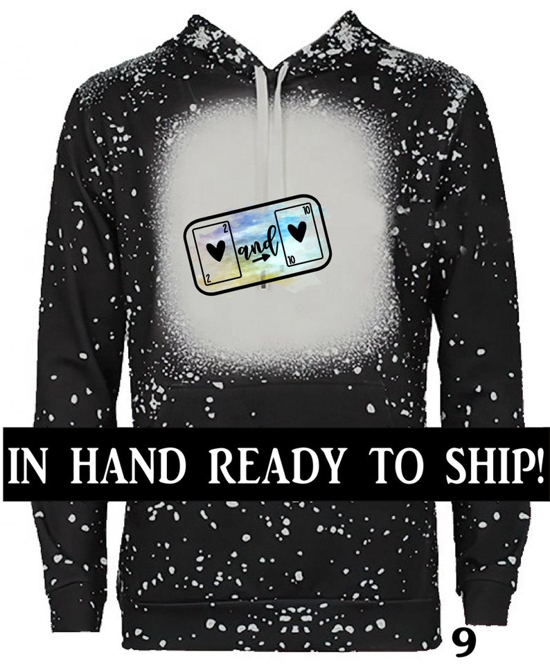 K&ouml;nnte beinhalten: Schwarzer Hoodie mit einem wei&szlig;en Spritzmuster. Der Hoodie hat eine Grafik mit zwei Spielkarten mit Herzen, den Zahlen 2 und 10 und dem Wort "and" dazwischen. Der Text "IN HAND READY TO SHIP!" befindet sich am unteren Rand des Bildes.