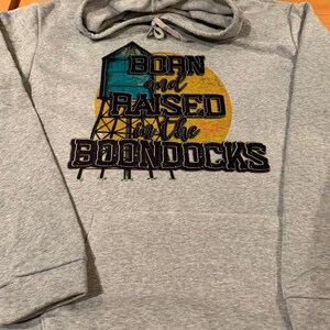 May include: A gray hoodie with a drawstring hood and a front pocket. The hoodie features a graphic of a water tower and the text "Born and Raised in the Boondocks" in a distressed font, with a yellow and orange sun in the background.