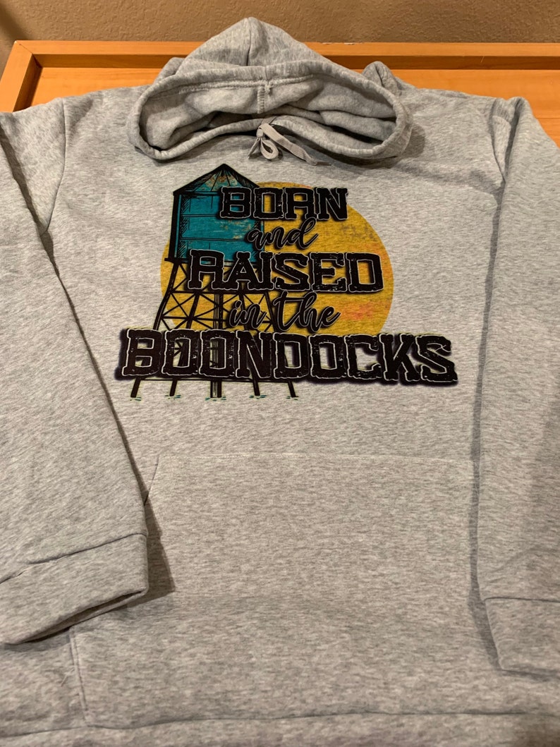 May include: A gray hoodie with a drawstring hood and a front pocket. The hoodie features a graphic of a water tower and the text "Born and Raised in the Boondocks" in a distressed font, with a yellow and orange sun in the background.