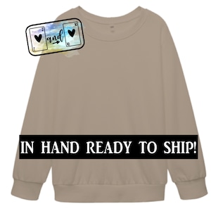 May include: A light brown sweatshirt with the text "IN HAND READY TO SHIP!" printed on the bottom. The sweatshirt has a graphic of two playing cards, a 2 of hearts and a 10 of hearts, with the word "and" and an arrow between them.