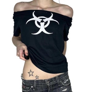 Può includere: Una t-shirt nera con spalle scoperte caratterizzata da un grande simbolo di rischio biologico bianco stampato sul petto. Il capo presenta maniche corte e un'ampia scollatura che poggia sulle spalle, abbinato a jeans in denim grigio scuro consumati.
