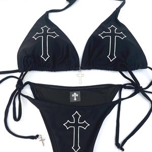 May include: A black bikini set featuring a white cross outline on each triangle cup and the centre of the bottom. The top includes a silver cross charm at the centre, while the side ties of the bottom are adorned with small silver cross charms.