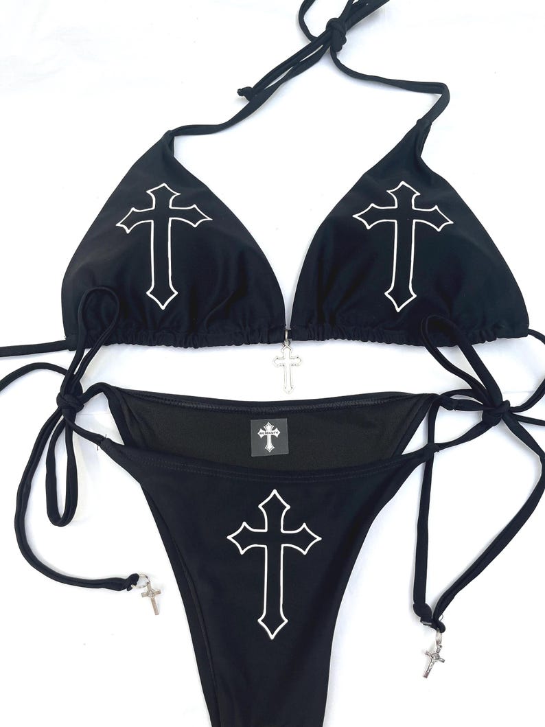May include: A black bikini set featuring a white cross outline on each triangle cup and the centre of the bottom. The top includes a silver cross charm at the centre, while the side ties of the bottom are adorned with small silver cross charms.