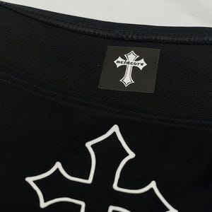 May include: A black garment featuring a large white outlined cross graphic on the front. A square black label is sewn near the neckline, displaying a white cross icon and the text acidcore in a stylised gothic font with white lettering.