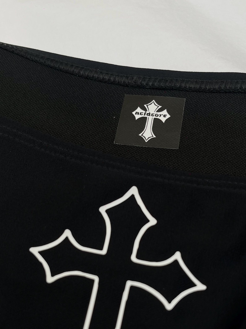 May include: A black garment featuring a large white outlined cross graphic on the front. A square black label is sewn near the neckline, displaying a white cross icon and the text acidcore in a stylised gothic font with white lettering.