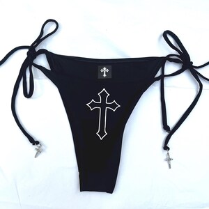 May include: A black bikini bottom featuring side tie closures with silver-toned cross charms attached to the ends. The front displays a large white outlined cross graphic in the centre and a small square label with a cross icon at the waistband.