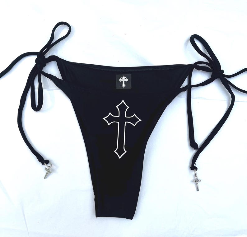 May include: A black bikini bottom featuring side tie closures with silver-toned cross charms attached to the ends. The front displays a large white outlined cross graphic in the centre and a small square label with a cross icon at the waistband.