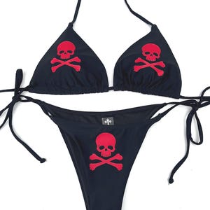 Può includere: Un set bikini nero con una grafica rossa a forma di teschio e ossa incrociate su ogni coppa triangolare e al centro dello slip. Il top presenta sottili laccetti da annodare al collo e sui lati, mentre lo slip ha laccetti laterali regolabili.