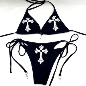 Könnte beinhalten: Ein schwarzer zweiteiliger Bikini mit weißen Kreuzmotiven auf dem Triangel-Oberteil und dem seitlich zu bindenden Unterteil. Der Bikini verfügt über dünne schwarze Bindebänder und kleine silberfarbene Kreuzanhänger am Oberteil und an den Bändern.