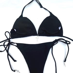 May include: A black bikini set featuring a triangle top with circular cutouts on each cup and a silver-toned cross charm at the centre. The matching bikini bottom has side ties, each finished with a small silver-toned cross charm at the ends.