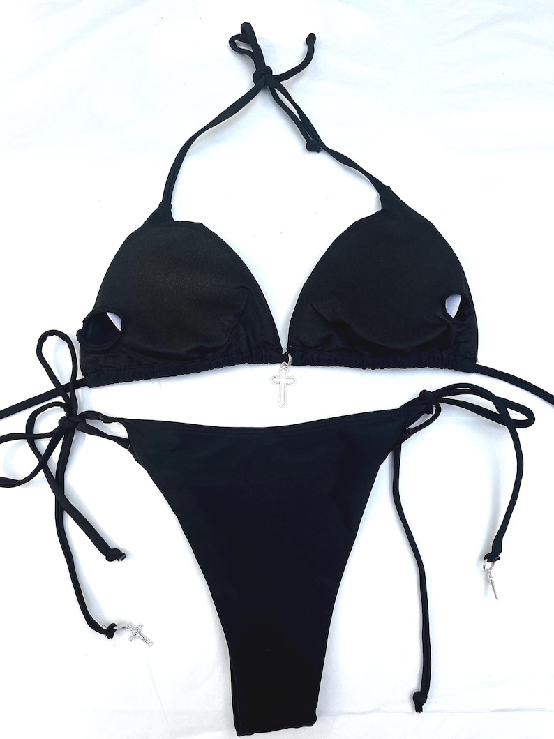 May include: A black bikini set featuring a triangle top with circular cutouts on each cup and a silver-toned cross charm at the centre. The matching bikini bottom has side ties, each finished with a small silver-toned cross charm at the ends.