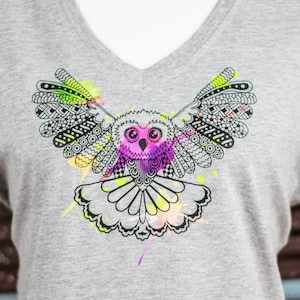Puede incluir: Una camiseta gris de cuello en V con un gráfico en blanco y negro de un búho con salpicaduras de pintura de colores. El búho tiene ojos rosas y un diseño detallado.