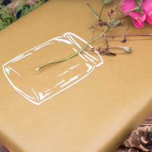 Puede incluir: Una bolsa de regalo de papel marrón con un contorno blanco de un frasco de vidrio. Una flor rosa con tallos verdes se coloca encima de la bolsa.