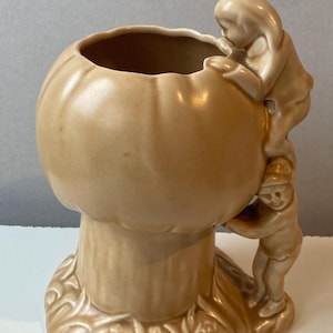 Peut inclure: Un vase en céramique de couleur beige en forme de champignon. Deux petites figurines grimpent sur le côté du vase. La base du vase présente un motif herbeux texturé.