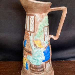 May include: A brown ceramic jug with a scene of a person sitting in a chair in front of a fireplace. The jug has a handle and a spout. The scene is painted in shades of brown, blue, green, and yellow.