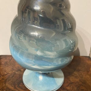 Pode incluir: Um vaso decorativo com um design bulboso e em camadas. O vaso tem um gradiente de cores, passando de um topo escuro e reflexivo para uma base azul clara. A base é um pedestal circular e o vaso tem um acabamento brilhante.
