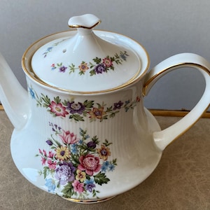 Puede incluir: Tetera de porcelana blanca con un diseño floral en rosa, morado, amarillo y azul. La tetera tiene un ribete dorado y una tapa a juego. El asa y el pico son curvos. Un artículo clásico para el servicio de té.