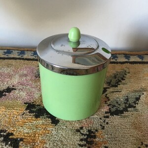 Peut inclure: Un pot à sucre en céramique vert avec un couvercle argenté et un petit bouton vert sur le dessus. Le bol est posé sur un tapis à motifs.