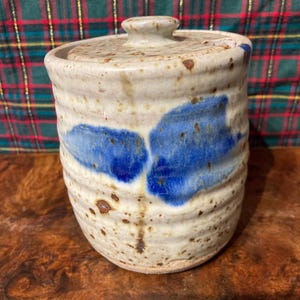 Puede incluir: Un tarro de cerámica con tapa, con un esmalte marrón moteado y detalles azules. El tarro tiene una forma redondeada y una abertura ligeramente acampanada.