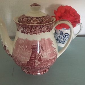 Könnte beinhalten: Eine rote und weiße Teekanne aus Keramik mit einem Blumendesign und einer Windmühlen-Szene. Die Teekanne hat einen Deckel und einen Ausguss.