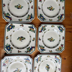 Może przedstawiać: Sześć kwadratowych ceramicznych talerzy z kwiatowym wzorem. Każdy talerz ma biały środek z małą kompozycją kwiatową i pierścieniem kwiatów na krawędzi. Talerze mają zieloną obwódkę i są ustawione na drewnianej powierzchni.