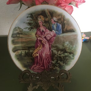 Könnte beinhalten: Ein Porzellanteller mit einer bemalten Szene eines Paares in einer Gartenlandschaft. Die Frau trägt ein rosa Kleid und der Mann einen blauen Anzug. Der Teller steht auf einem goldenen Metallständer.