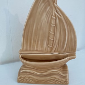 Peut inclure: Un pot de fleurs en céramique beige en forme de voilier. Le pot a une grande voile avec des lignes verticales et une petite base en forme de bateau. La base est texturée pour ressembler à des vagues. C'est un objet décoratif.