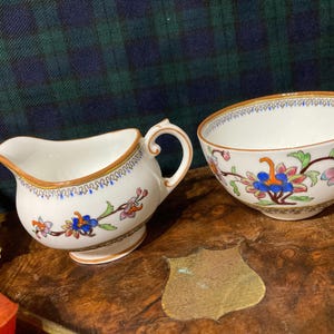 Könnte beinhalten: Ein weißes Porzellan-Sahnegießer- und Zuckergefäß-Set mit einem Blumenmuster in Blautönen, Rosa, Grün und Gold. Das Set verfügt über ein zartes Blumenmuster mit goldener Verzierung.