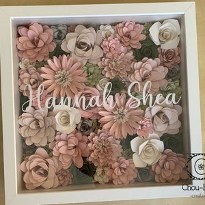 May include: A white shadow box with a floral arrangement of pink and green paper flowers. The name "Hannah Shea" is written in white lettering on the front of the box.
