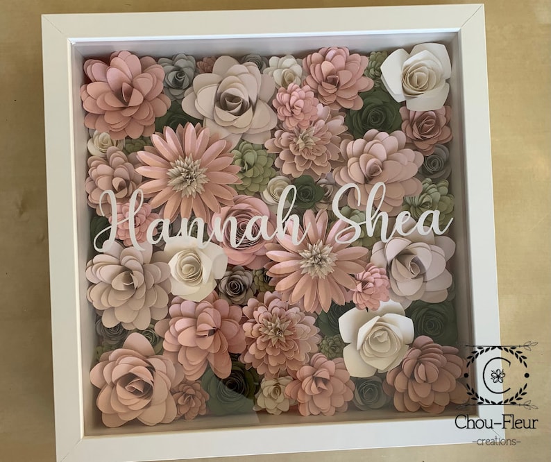 May include: A white shadow box with a floral arrangement of pink and green paper flowers. The name "Hannah Shea" is written in white lettering on the front of the box.