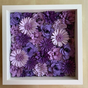 May include: A white frame with a purple and lavender paper flower arrangement. The flowers are arranged in a square shape and are all different sizes and shapes. The flowers are made of paper and have a realistic look.