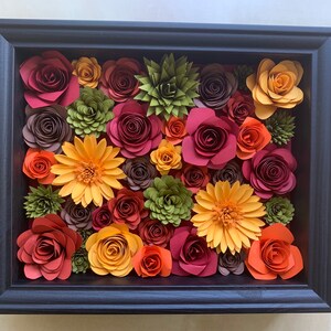 May include: A framed paper flower arrangement with a variety of colors, including red, orange, yellow, and green. The flowers are arranged in a circular pattern and are surrounded by a black frame.